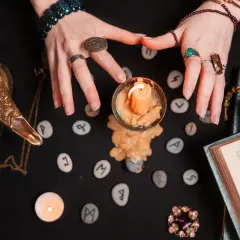 Dłonie nad runami i płonącą świecą. W tle lampa, książka i biżuteria. Czas na analizę 4 roku numerologicznego.