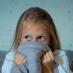 Przerażona dziewczynka z niebieskimi oczami chowa się pod kołdrą, zastanawiając się, czy dzieci widzą duchy.