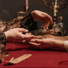 Dwie kobiety przy stole, jedna czyta z dłoni drugiej. W tle świece i butelki, atmosfera tajemnicza, jak w domu astrologii.