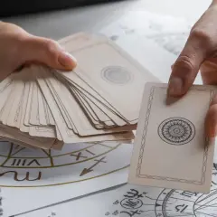 Dwie dłonie trzymają talię kart, jedna wybiera kartę z ozdobnym wzorem. W tle widać koło zodiaku, sugerujące wróżenie z kart tarot.