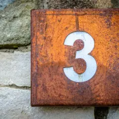 Zardzewiała tabliczka z numerem 3, symbolizująca numerologię 3, przyczepiona do kamiennej ściany.