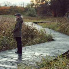Kobieta w jesiennej kurtce na drewnianej ścieżce, otoczona trzcinami. Chwila spokoju sprzyja samouzdrawianiu i medytacji.