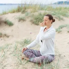 Kobieta siedzi na plaży, wykonując ćwiczenie oddechowe. To przykład pranajamy, czyli kontroli oddechu w jodze.