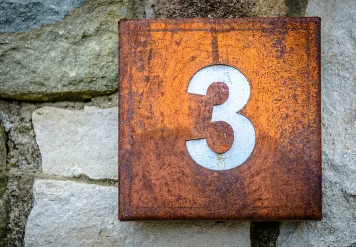 Zardzewiała tabliczka z numerem 3, symbolizująca numerologię 3, przyczepiona do kamiennej ściany.