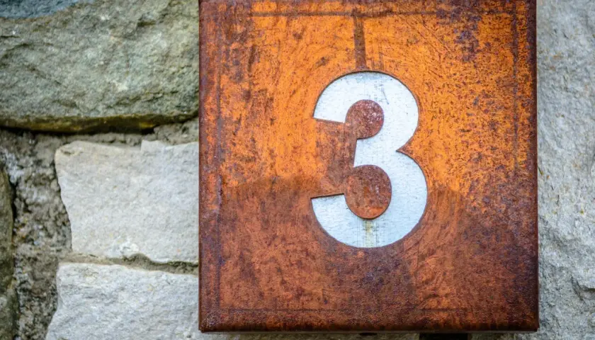 Zardzewiała tabliczka z numerem 3, symbolizująca numerologię 3, przyczepiona do kamiennej ściany.