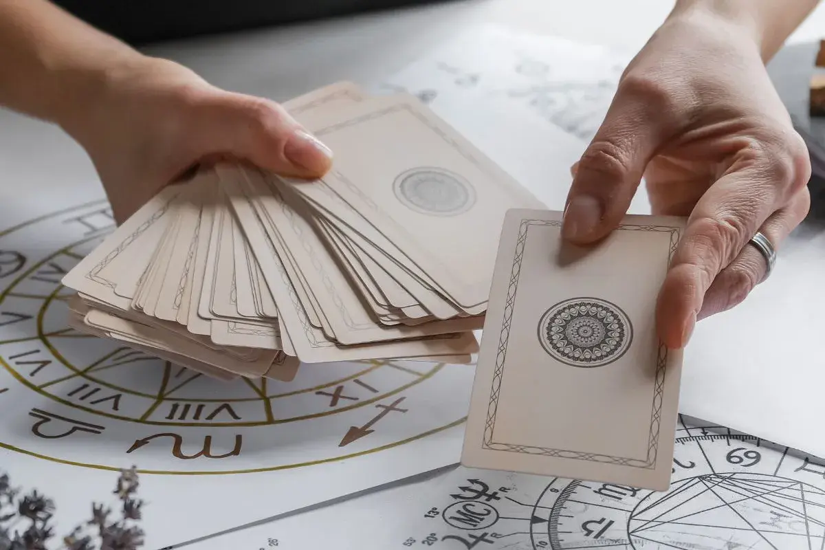 Dwie dłonie trzymają talię kart, jedna wybiera kartę z ozdobnym wzorem. W tle widać koło zodiaku, sugerujące wróżenie z kart tarot.