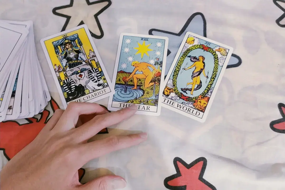 Dłoń wskazuje na karty Tarota: Rydwan, Gwiazda i Świat. Wygląda na to, że ktoś wybiera swoje 2 monety tarot.