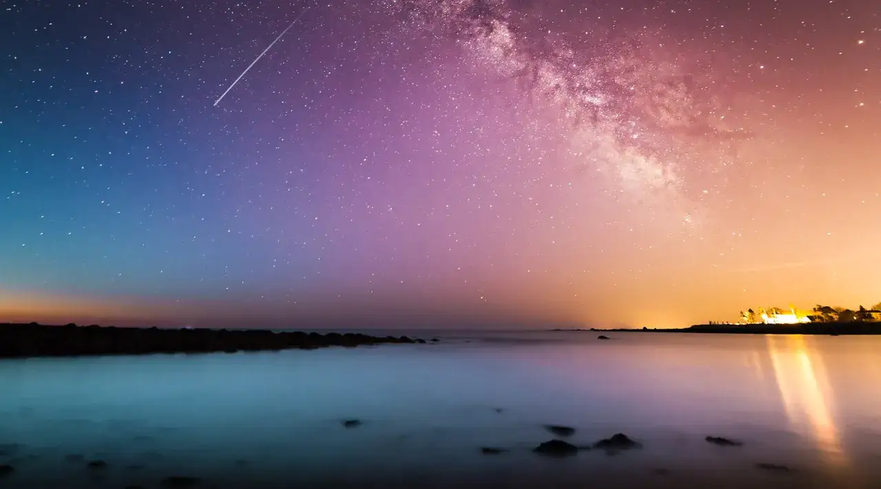 Gwieździste niebo nad spokojnym morzem, kiedy jest przesilenie zimowe. Spadająca gwiazda przecina fioletowo-pomarańczowy firmament.